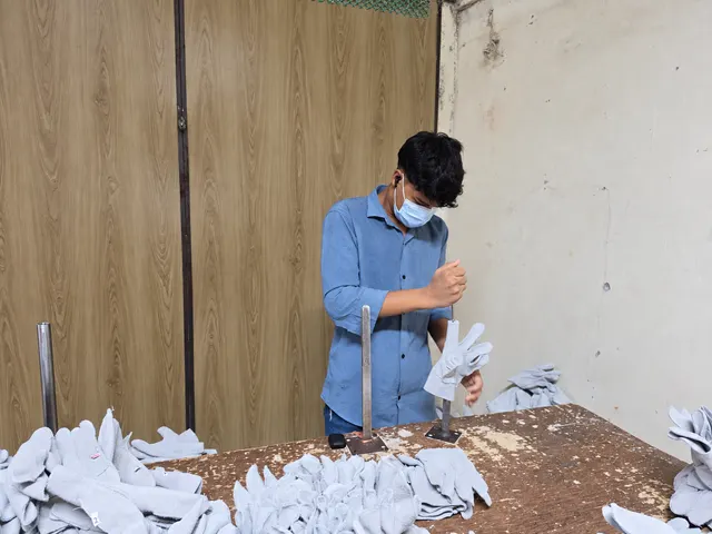 Masked team member in a blue shirt working on a leather glove at a metal form